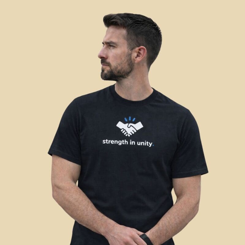 Man wearing a black MMH Statement Apparel T-shirt featuring the white handshake icon with blue accent and the slogan “strength in unity.” on the front, clean minimalist design on a neutral background.