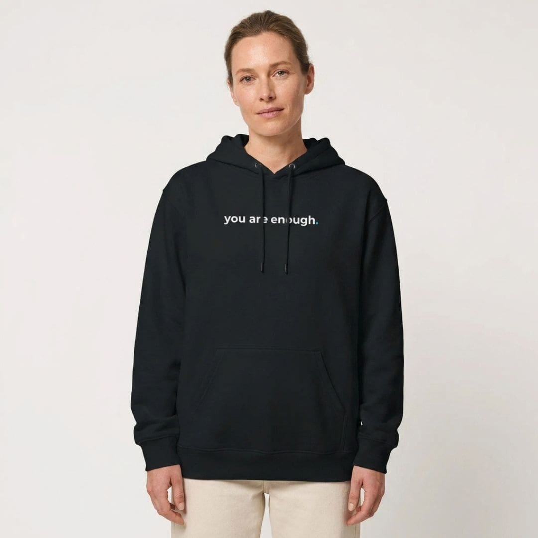 A woman stands against a plain, light-coloured background wearing a black hoodie with the words "you are enough." printed across the chest in white text. She pairs it with light beige trousers, creating a clean and understated look. The overall style is minimal and calm, with the slogan serving as the focal point of the image.