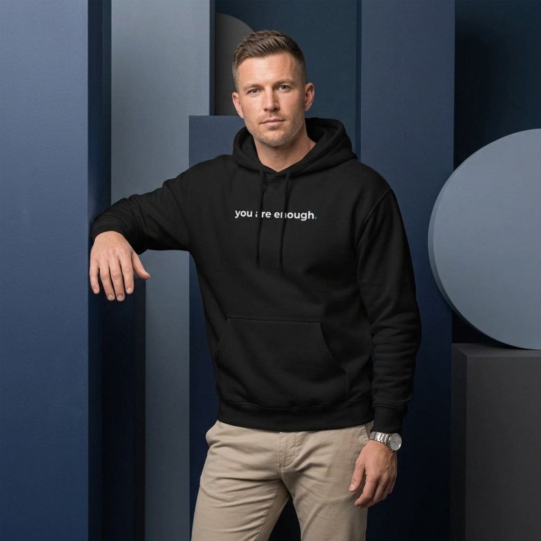 A man stands against a backdrop of geometric blue shapes, wearing a black hoodie printed with the words "you are enough." He pairs the hoodie with beige trousers and a silver wristwatch, creating a smart-casual look. The clean, modern setting and neutral tones draw attention to the motivational message on his clothing.
