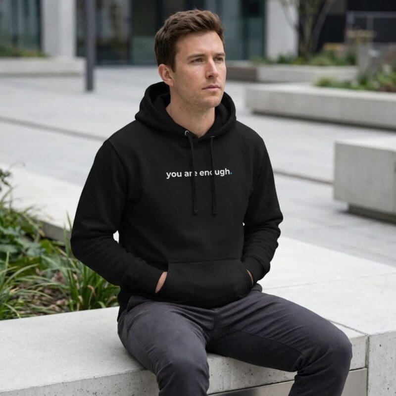 A man is sitting on a concrete bench in a modern outdoor setting, with greenery and contemporary buildings in the background. He is wearing a black hoodie with the words "you are enough." printed in white across the front, paired with dark trousers. His hands are in his pockets as he gazes thoughtfully into the distance.