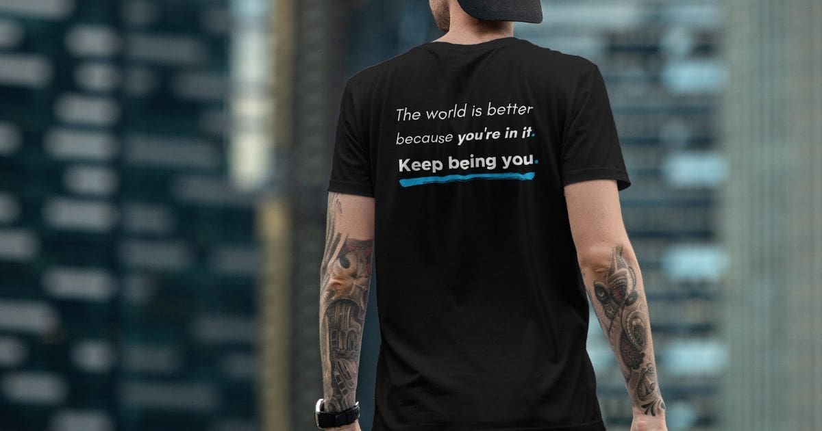 Back view of a man wearing a black statement T-shirt with the message “The world is better because you’re in it. Keep being you.” in an urban city setting.