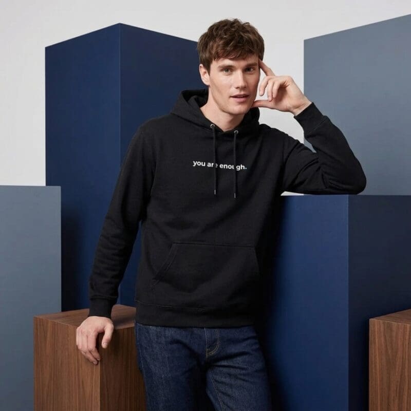 Male model wearing a black MMH Clothing “You Are Enough” premium hoodie in a studio setting with blue geometric blocks. The front chest print reads “you are enough.” in white with the hoodie drawstrings partly covering the text.