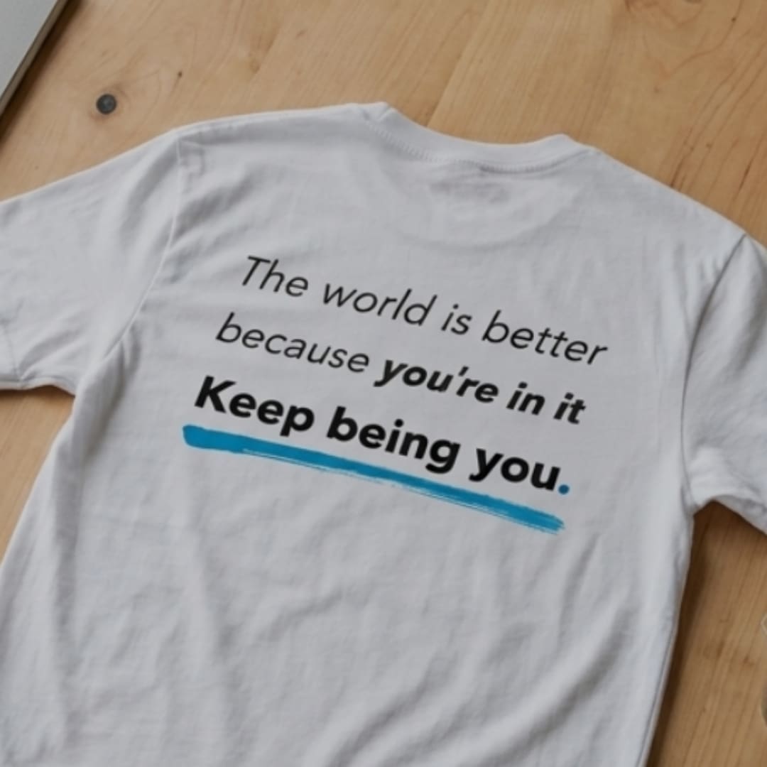 White You Are Enough T-shirt laid flat on a wooden surface, showing the back print: “The world is better because you’re in it. Keep being you.” with a blue underline.